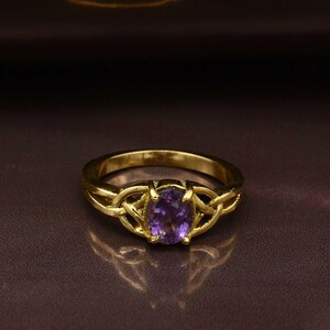 Pode incluir: Um anel dourado com uma pedra preciosa oval roxa. O anel tem um intrincado design de nó celta na banda, com a pedra preciosa no centro. O anel é exibido em uma superfície marrom avermelhada escura.