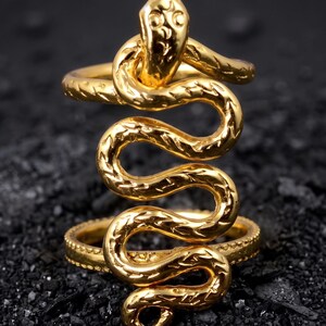 May include: A gold-toned ring featuring a coiled snake design that wraps around the finger. The snake has a textured, scale-like pattern along its body and a defined head at the top. The band of the ring is thin and circular, supporting the vertical snake figure.