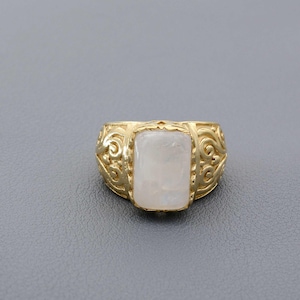 Puede incluir: Un anillo dorado con un diseño ornamental y ondulado en la banda. El anillo presenta una piedra rectangular de color claro en el centro. El anillo está sobre un fondo gris.