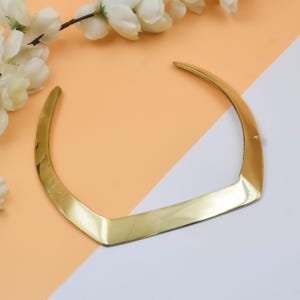May include: A gold-colored collar necklace with a V-shaped design. The necklace has a smooth, polished finish and is set against a peach and white background. White flowers are in the upper left corner.