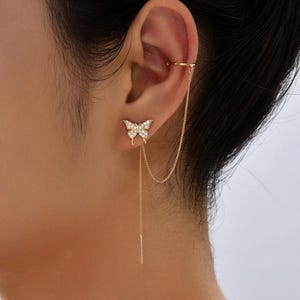 Könnte beinhalten: Goldfarbenes Schmetterling-Ohr-Set mit Ohrklemme. Der Schmetterling-Ohrstecker ist mit kleinen, klaren Kristallen verziert. Eine zarte Kette verbindet den Ohrring mit einer Manschette, die sich um das Ohr legt. Der Ohrring hat einen langen, baumelnden Stab.