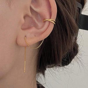 May include: A close-up of a gold ear cuff and a gold chain earring. The ear cuff has a double-band design. The earring features a delicate chain that dangles down with a small bar at the end. The jewelry is worn on a person's ear.