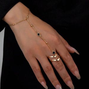 Könnte beinhalten: Goldfarbenes Handkettenarmband mit schwarzen kreisförmigen Akzenten und einem doppelten Chevron-Ring. Das Armband ist durch eine zarte Kette mit dem Ring verbunden, was einen stilvollen und modernen Look erzeugt. Der Schmuck wird auf einer Hand mit langen, polierten Nägeln präsentiert.