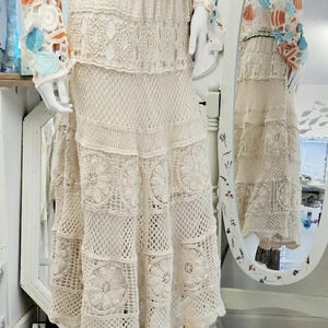 May include: A cream crochet skirt and top set on a mannequin. The top has seashell appliqués in orange, blue, and white. The skirt features a tiered design with floral and geometric patterns. A seashell belt and white sandals complete the outfit.