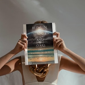 Könnte beinhalten: Eine Person hält ein Buch mit dem Titel „Between Shores and Stars“ mit einem Bild der Erde und des Ozeans. Eine große goldene Kette ist sichtbar. Die Person trägt ein weißes Strickoberteil.