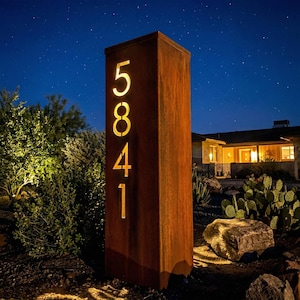 May include: A tall, rectangular address sign made of weathered metal. The numbers "5841" are cut out, allowing light to shine through. The sign is set in a landscaped yard at night, with a house in the background.