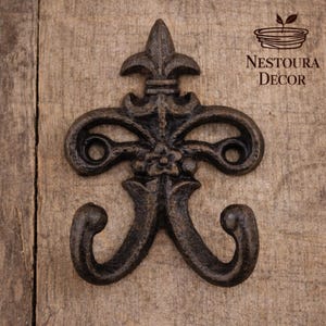 May include: Ornate, dark brown metal wall hook with a fleur-de-lis design. The hook has two curved arms and a decorative floral element. The logo "Nestoura Decor" is visible in the upper right corner.