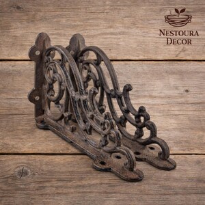 May include: Two ornate, cast iron shelf brackets with a distressed, rust-colored finish. The brackets feature intricate scrollwork and decorative detailing, perfect for adding a vintage touch to any space. The brackets are shown on a wooden surface.