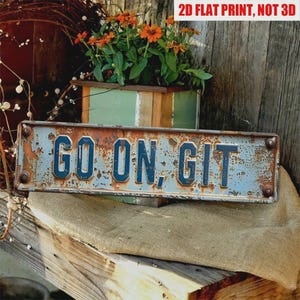 May include: A rectangular, weathered metal sign with the words "GO ON, GIT" in blue lettering. The sign has a distressed, rusty appearance, mounted on a wooden surface with a burlap cloth. The background includes a planter box with orange flowers.