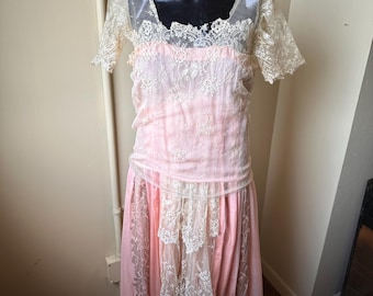 May include: A vintage pink dress with a square neckline and short sleeves. The dress features delicate white lace detailing on the bodice, sleeves, and skirt. The skirt has a layered design with a flowing, romantic silhouette. The dress is displayed on a black mannequin.