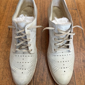 May include: A pair of off-white leather oxford shoes featuring a lace-up closure with matching laces, a rounded toe, and decorative perforated brogue detailing across the vamp. The interior of each shoe contains white tissue paper stuffing.