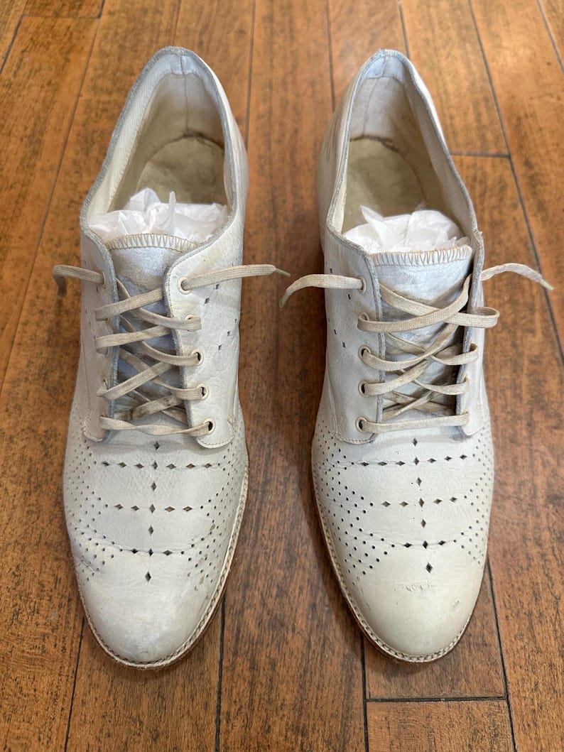 May include: A pair of off-white leather oxford shoes featuring a lace-up closure with matching laces, a rounded toe, and decorative perforated brogue detailing across the vamp. The interior of each shoe contains white tissue paper stuffing.