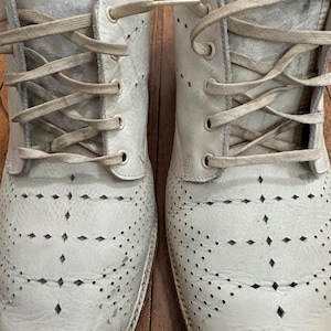 May include: A pair of off-white leather oxford shoes featuring a decorative diamond-shaped perforated pattern across the toe box and vamp. The shoes include matching off-white flat laces threaded through metal eyelets and a textured leather finish.