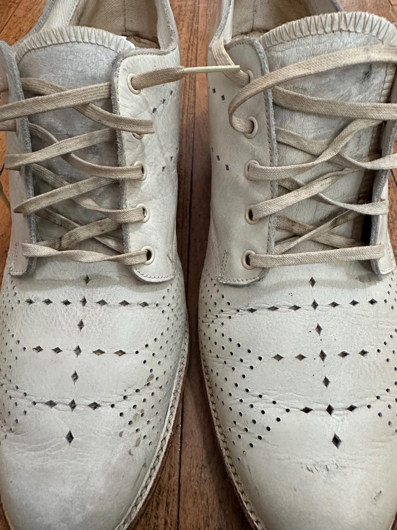 May include: A pair of off-white leather oxford shoes featuring a decorative diamond-shaped perforated pattern across the toe box and vamp. The shoes include matching off-white flat laces threaded through metal eyelets and a textured leather finish.
