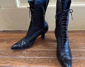May include: A pair of black leather lace-up boots featuring a pointed toe, a mid-calf height shaft, and a curved kitten heel. The boots have black laces threaded through metal eyelets that extend from the vamp to the top of the boot shaft.