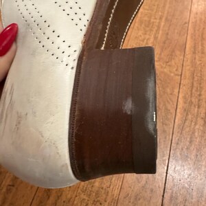 May include: A white leather oxford shoe featuring a perforated brogue pattern on the side and a dark brown stacked wooden heel. The shoe has a rounded toe and a sturdy construction with visible stitching along the edge of the dark brown sole.