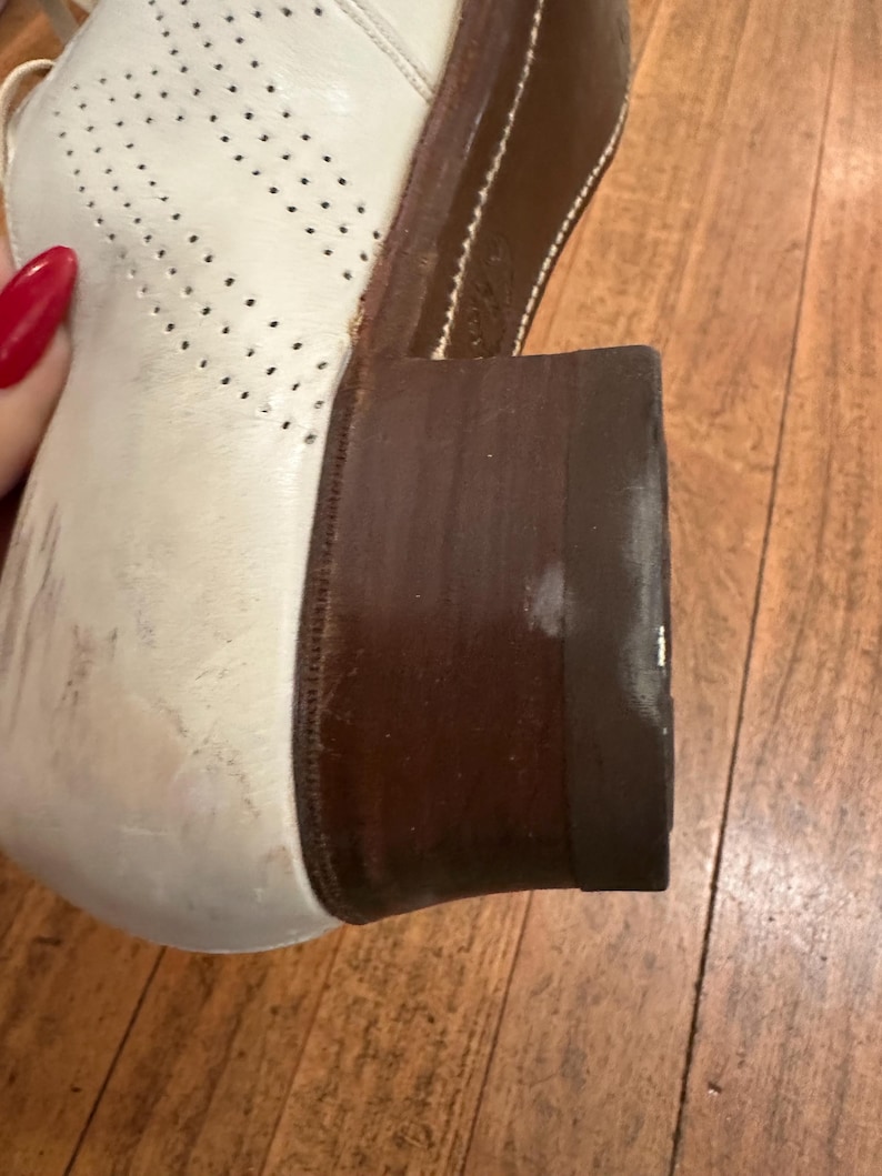 May include: A white leather oxford shoe featuring a perforated brogue pattern on the side and a dark brown stacked wooden heel. The shoe has a rounded toe and a sturdy construction with visible stitching along the edge of the dark brown sole.