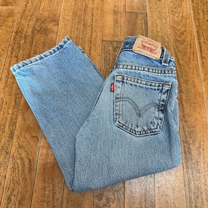 May include: Light blue denim jeans featuring a classic five-pocket design, a tan leather-like patch on the back waistband with Levi Strauss & Co. branding, and a small red tab on the back pocket with the Levi's logo in white lettering.