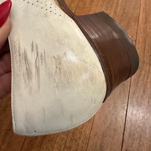 May include: A white leather shoe featuring a perforated pattern on the upper section, a stacked brown wooden heel, and visible scuff marks on the side of the heel counter. The shoe displays signs of wear with dark streaks across the white surface.
