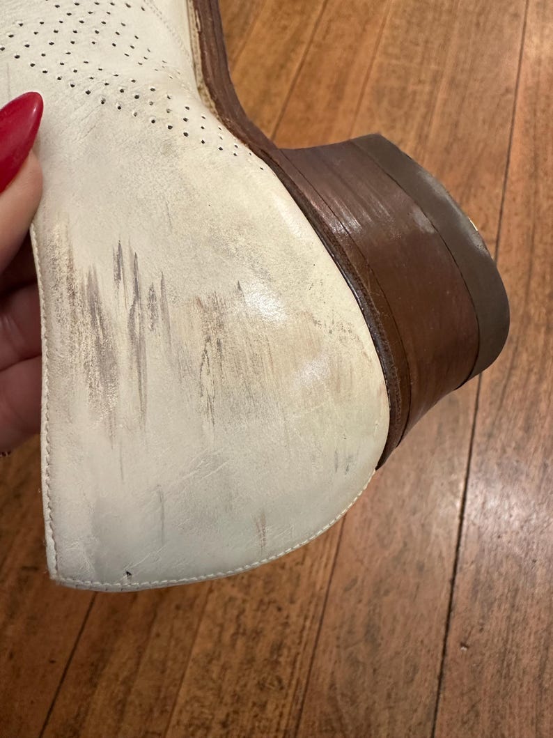 May include: A white leather shoe featuring a perforated pattern on the upper section, a stacked brown wooden heel, and visible scuff marks on the side of the heel counter. The shoe displays signs of wear with dark streaks across the white surface.