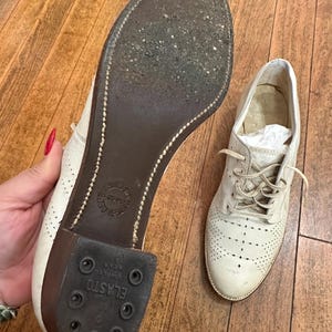 May include: A pair of off-white leather oxford shoes with decorative perforations on the toe and sides. One shoe shows the dark brown sole with a visible Elasto heel and stitched detailing, while the other shoe is shown from the top with laces.