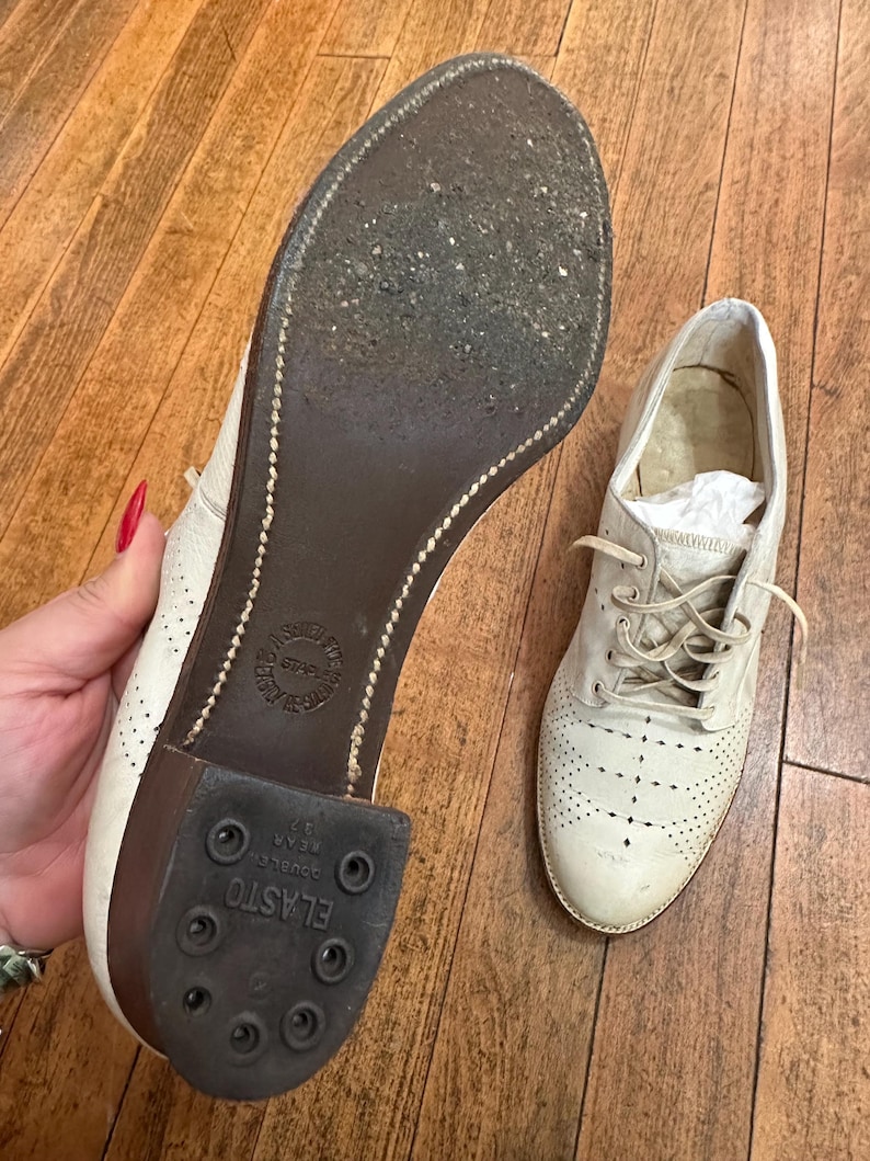 May include: A pair of off-white leather oxford shoes with decorative perforations on the toe and sides. One shoe shows the dark brown sole with a visible Elasto heel and stitched detailing, while the other shoe is shown from the top with laces.