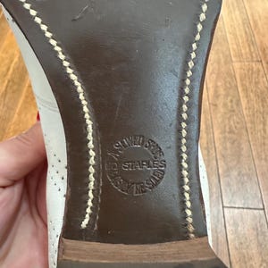 May include: The dark brown leather sole of a white shoe featuring visible light-coloured stitching along the edges. A circular stamp is embossed in the centre of the sole with the text NO STAPLES, A SEWED SOLE, and EASILY RE-SOLED.