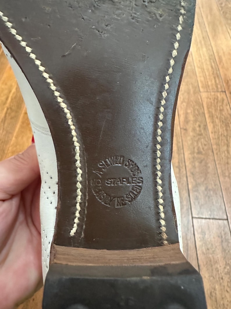 May include: The dark brown leather sole of a white shoe featuring visible light-coloured stitching along the edges. A circular stamp is embossed in the centre of the sole with the text NO STAPLES, A SEWED SOLE, and EASILY RE-SOLED.