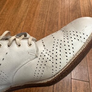 May include: A white leather oxford shoe featuring decorative brogue perforations across the toe and side panels. The shoe includes matching white laces, a rounded toe, and a brown stacked heel with a visible welt construction.