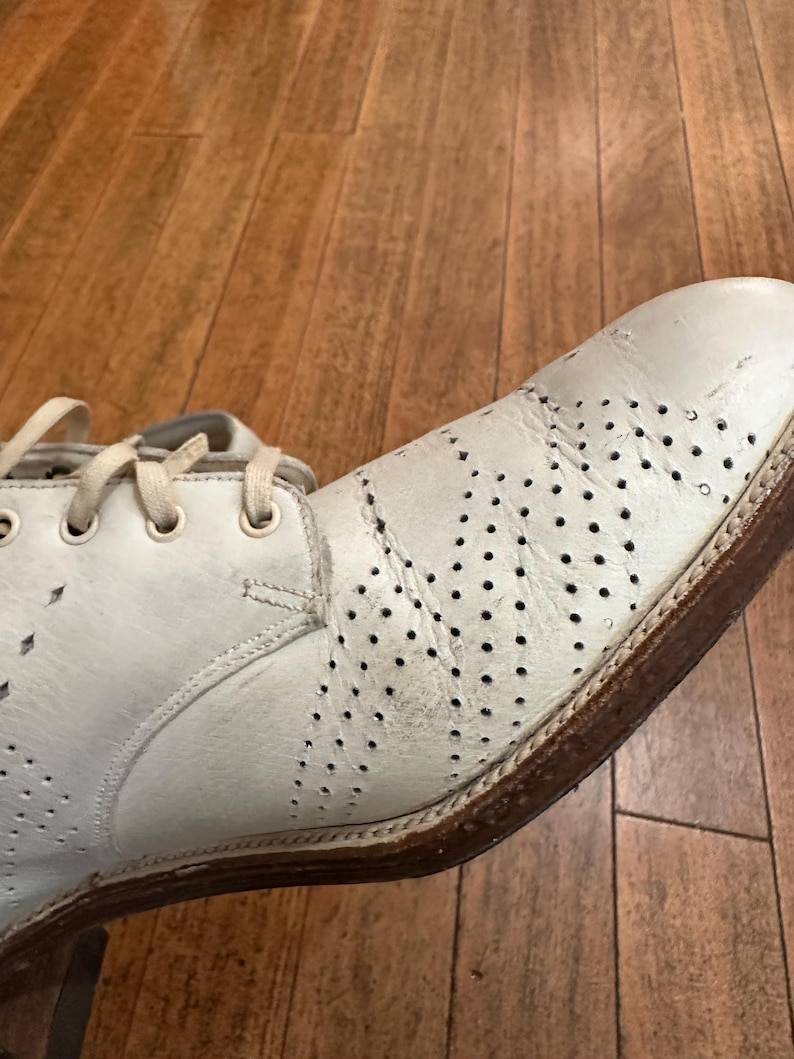 May include: A white leather oxford shoe featuring decorative brogue perforations across the toe and side panels. The shoe includes matching white laces, a rounded toe, and a brown stacked heel with a visible welt construction.