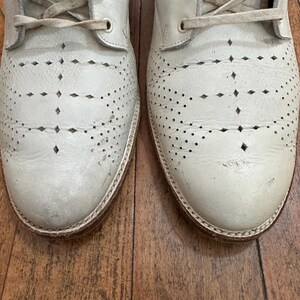 May include: A pair of off-white leather lace-up boots featuring decorative brogue-style perforations and diamond-shaped cutouts across the toe and vamp. The boots have matching off-white laces and a visible stitched welt along the sole edge.