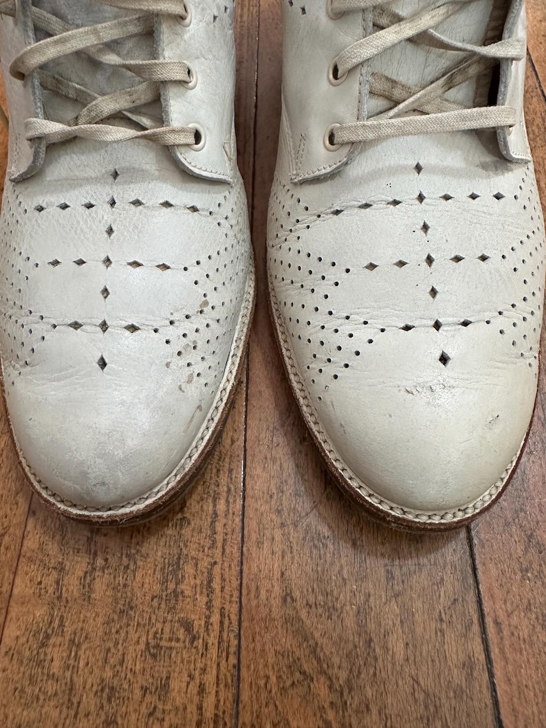 May include: A pair of off-white leather lace-up boots featuring decorative brogue-style perforations and diamond-shaped cutouts across the toe and vamp. The boots have matching off-white laces and a visible stitched welt along the sole edge.