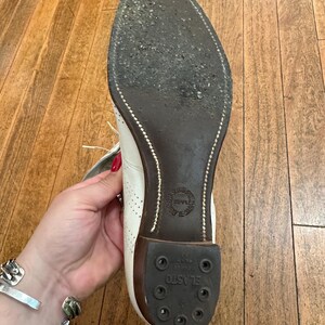 May include: A white shoe with a dark brown sole and a black rubber heel piece labelled ELASTO. The sole features visible stitching along the edge and a circular stamp in the centre. The upper part of the shoe shows white leather with perforations.