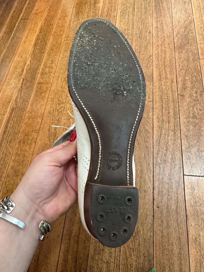 May include: A white shoe with a dark brown sole and a black rubber heel piece labelled ELASTO. The sole features visible stitching along the edge and a circular stamp in the centre. The upper part of the shoe shows white leather with perforations.