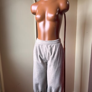 May include: Light gray, cropped sweatpants with a gathered elastic hem, displayed on a mannequin. The pants have a high waist and a loose fit through the legs. The mannequin is a bronze color and is on a brown stand.