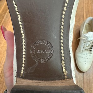 May include: The underside of a white shoe with a dark brown sole featuring visible light-coloured stitching along the edges. A circular stamp on the sole reads A SEWED SHOE NO STAPLES USED IN SOLE. A second white shoe is visible in the background.