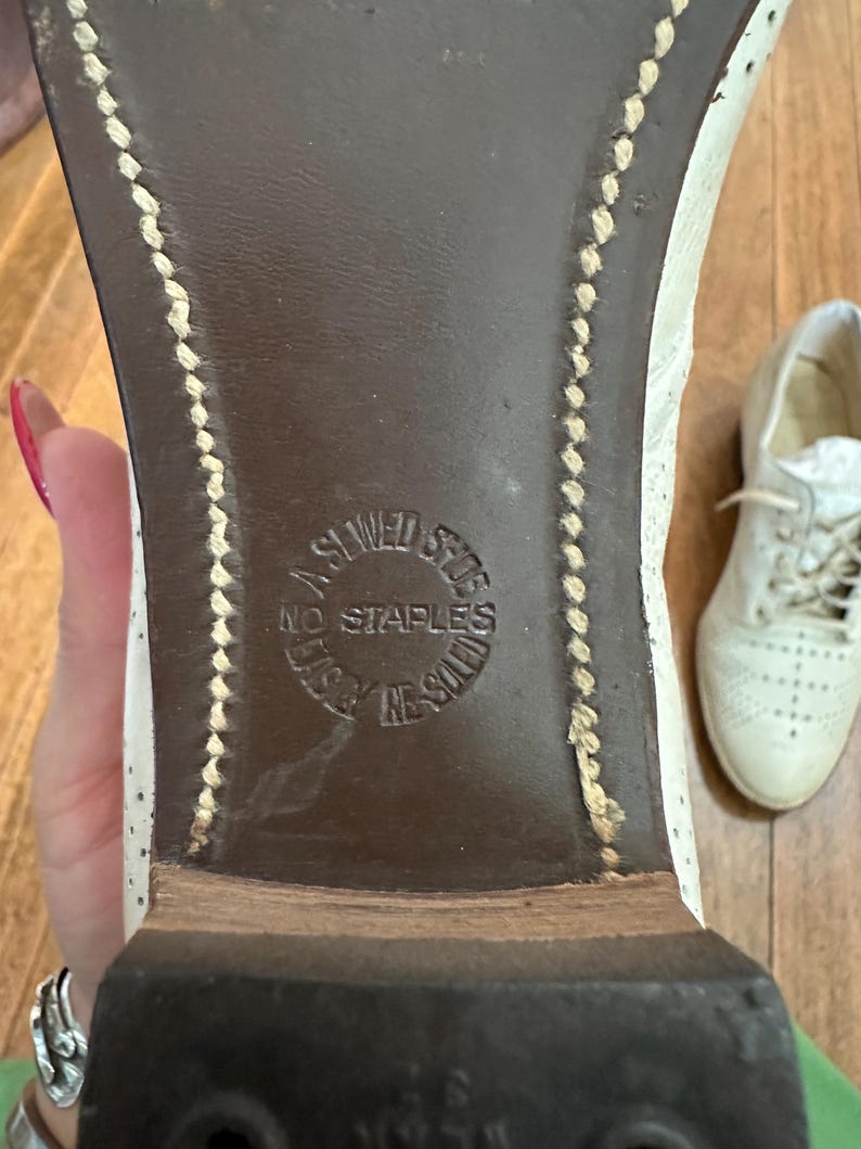 May include: The underside of a white shoe with a dark brown sole featuring visible light-coloured stitching along the edges. A circular stamp on the sole reads A SEWED SHOE NO STAPLES USED IN SOLE. A second white shoe is visible in the background.