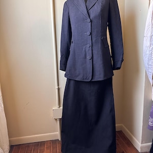 Pode incluir: Um fato azul marinho com riscas finas, composto por um blazer de manga comprida com três botões e uma saia comprida. O fato é exibido num manequim. O blazer tem um corte sob medida com lapela entalhada.