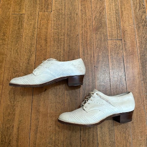 May include: A pair of off-white leather oxford shoes featuring decorative brogue perforations on the toe cap and sides, lace-up closures, and a low, stacked brown heel. The shoes have a rounded toe design and a smooth, light-coloured finish.