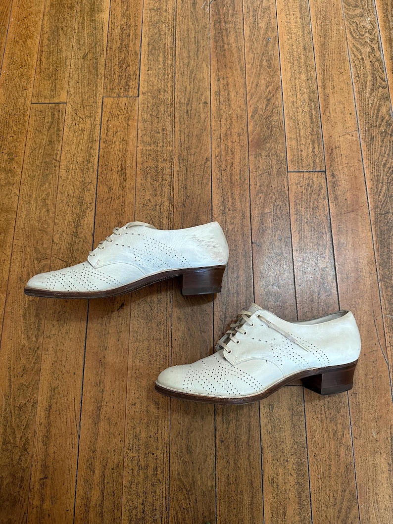 May include: A pair of off-white leather oxford shoes featuring decorative brogue perforations on the toe cap and sides, lace-up closures, and a low, stacked brown heel. The shoes have a rounded toe design and a smooth, light-coloured finish.