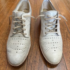 May include: A pair of off-white leather oxford shoes featuring a lace-up closure, a rounded toe, and intricate perforated brogue detailing across the vamp. The shoes have a low heel, a tan leather sole, and matching off-white laces.