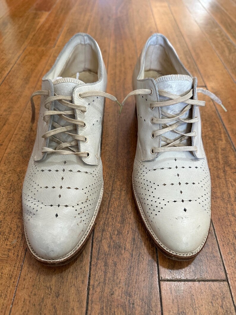 May include: A pair of off-white leather oxford shoes featuring a lace-up closure, a rounded toe, and intricate perforated brogue detailing across the vamp. The shoes have a low heel, a tan leather sole, and matching off-white laces.