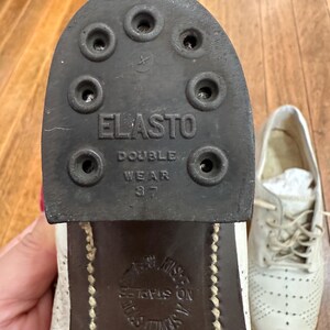 May include: A white leather shoe with a dark brown heel and sole. The heel features a black rubber tap labelled ELASTO DOUBLE WEAR 37 with six circular nail holes. The sole is embossed with the text NO STAPLES A SEWED SOLE.