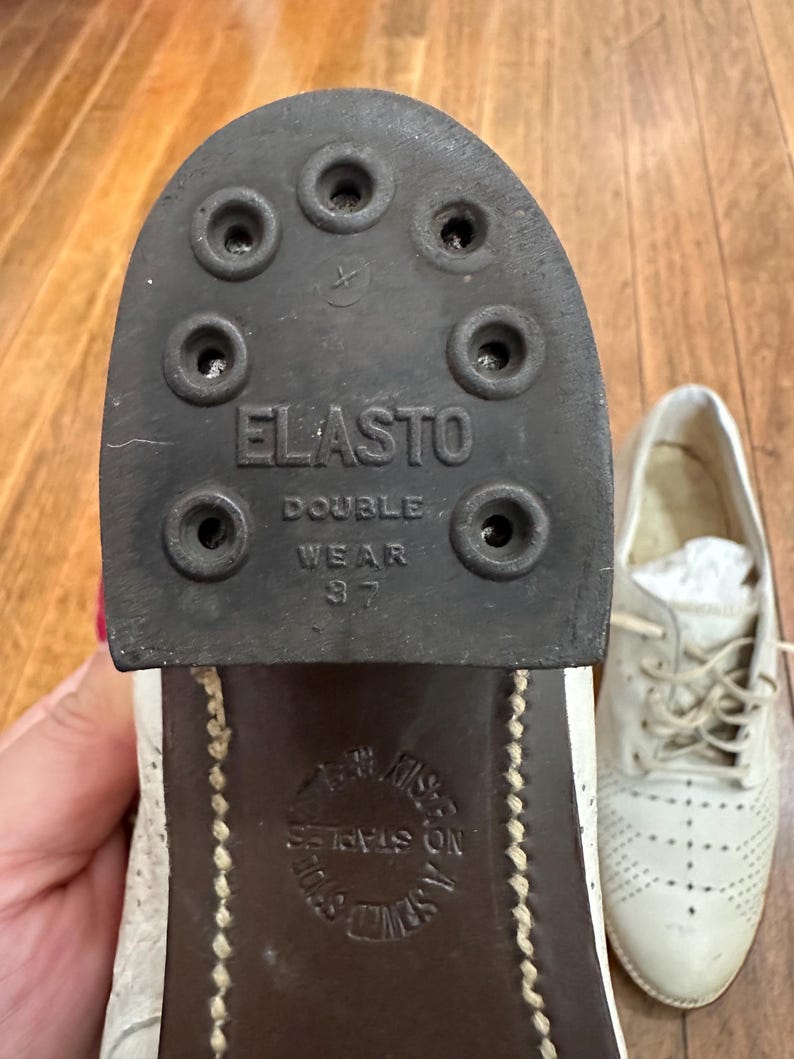 May include: A white leather shoe with a dark brown heel and sole. The heel features a black rubber tap labelled ELASTO DOUBLE WEAR 37 with six circular nail holes. The sole is embossed with the text NO STAPLES A SEWED SOLE.
