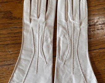 May include: A pair of off-white leather gloves. The gloves have a classic design with detailed stitching along the seams and fingers. The gloves are displayed on a wooden surface.