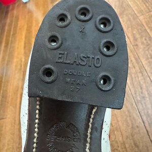 May include: A black rubber shoe heel with the text ELASTO DOUBLE WEAR 37 embossed on the surface. The heel features eight circular holes for attachment. Below the heel, a circular stamp on the shoe sole reads NO STAPLES and JOHN WHITE.