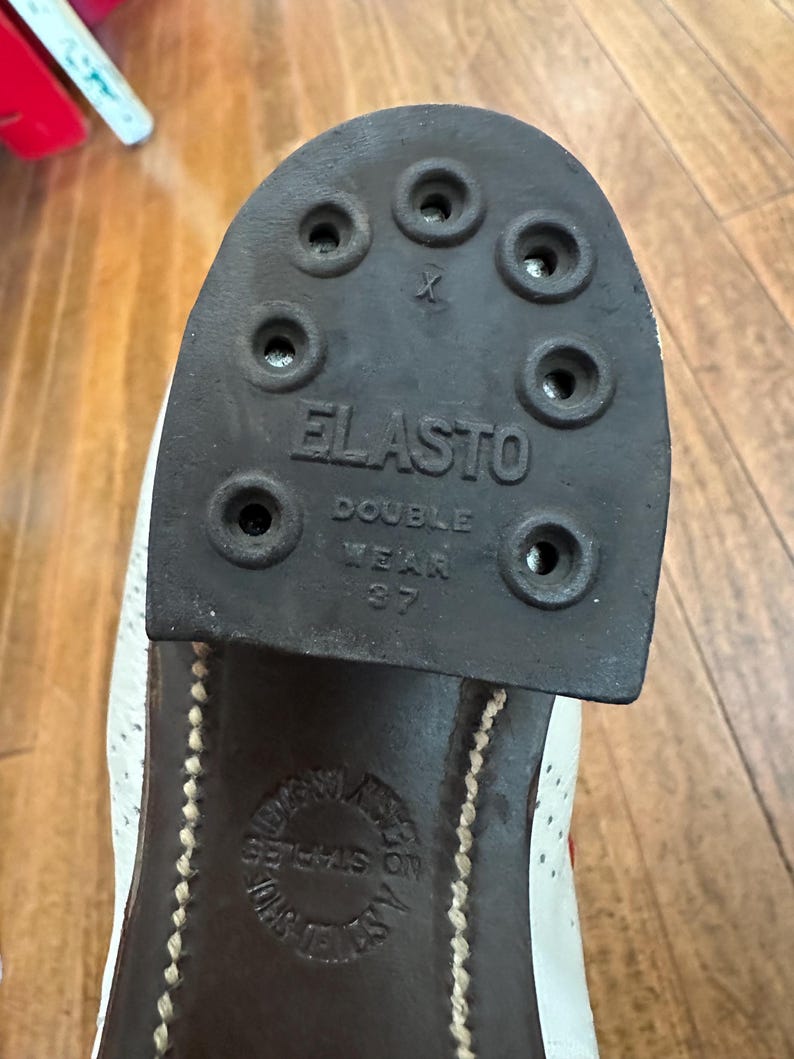 May include: A black rubber shoe heel with the text ELASTO DOUBLE WEAR 37 embossed on the surface. The heel features eight circular holes for attachment. Below the heel, a circular stamp on the shoe sole reads NO STAPLES and JOHN WHITE.