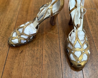May include: A pair of gold-toned T-strap heels featuring a cage-style cutout design on the toe box, a buckled ankle strap, and a mid-height heel. The interior of the shoes is lined in white, and the footbed displays the brand name Palter De Liso.