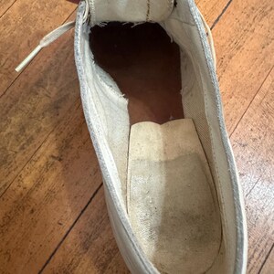 May include: A high-top trainer interior showing a white canvas tongue and lining, a dark brown heel section, and a light beige insole. The shoe features a visible lace eyelet and a worn, textured fabric construction throughout the inner cavity.