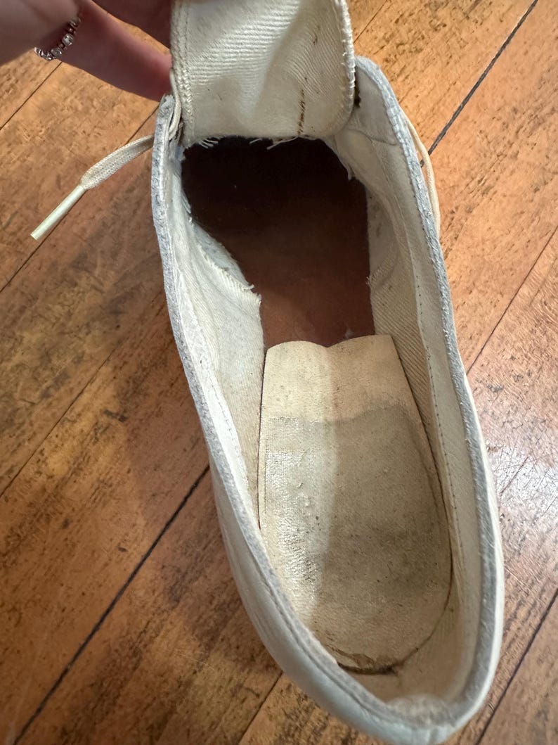May include: A high-top trainer interior showing a white canvas tongue and lining, a dark brown heel section, and a light beige insole. The shoe features a visible lace eyelet and a worn, textured fabric construction throughout the inner cavity.